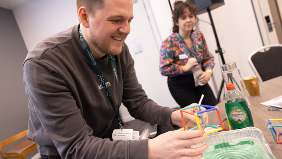 A person holds a cube made of straws above a tray filled with liquid. Another person looks on