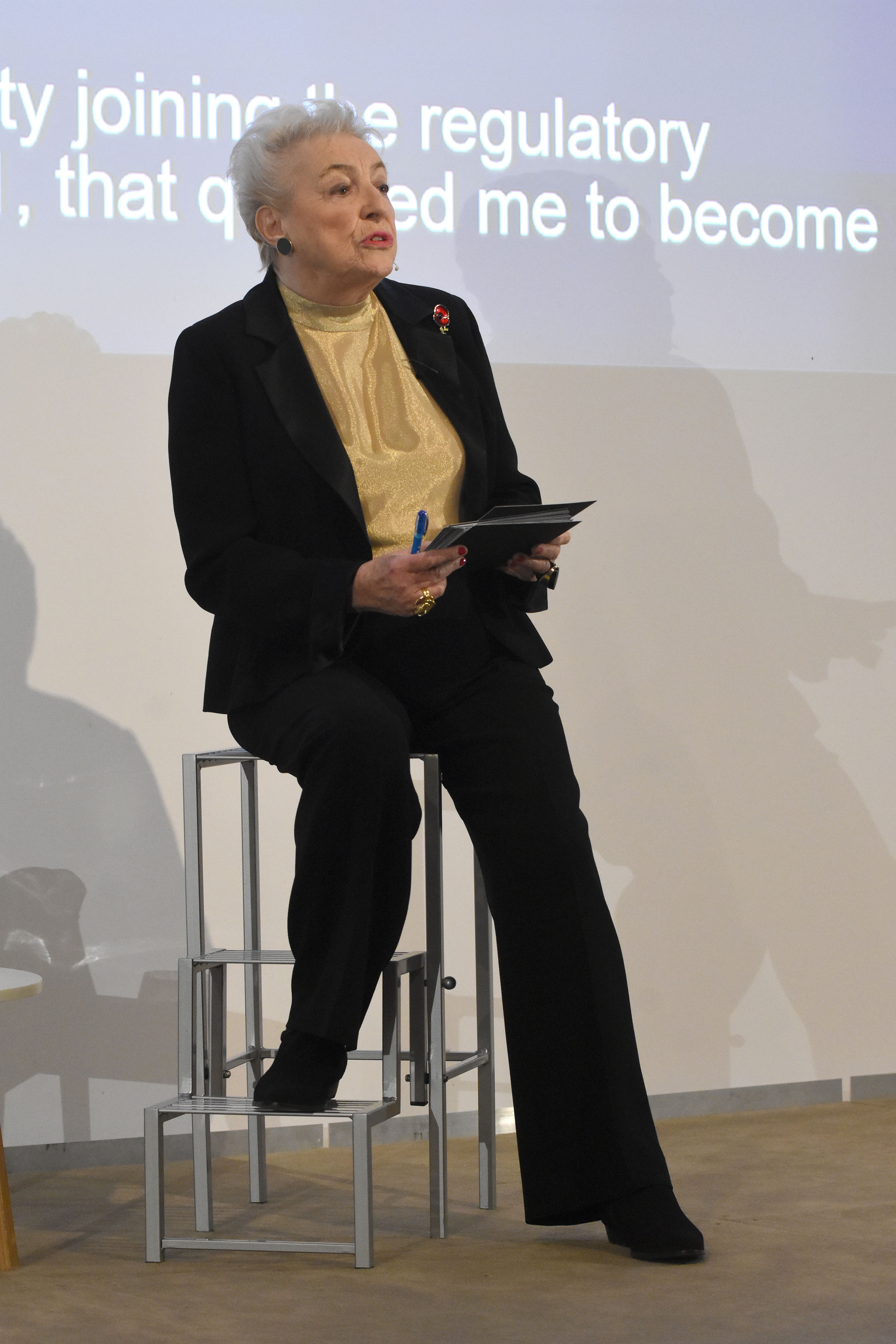 A woman in a suit, perched on a stool, giving a talk in front of a projector screen