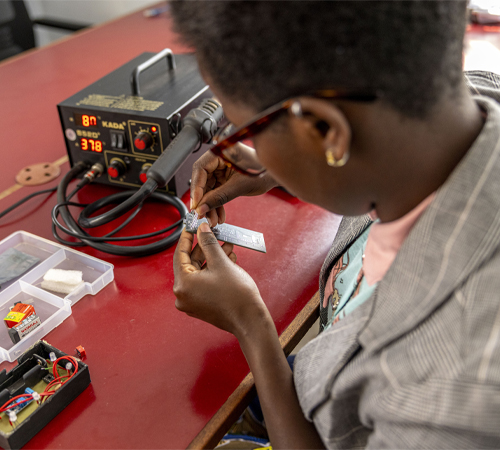 A young, Black woman, wearing glasses and a bright coloured tshirt under a light brown blazer, assembles a circuit board in an office while sat at a red table. There is black soldering station on the table in the background, and a tray of circuit parts and soldering materials in front of her.