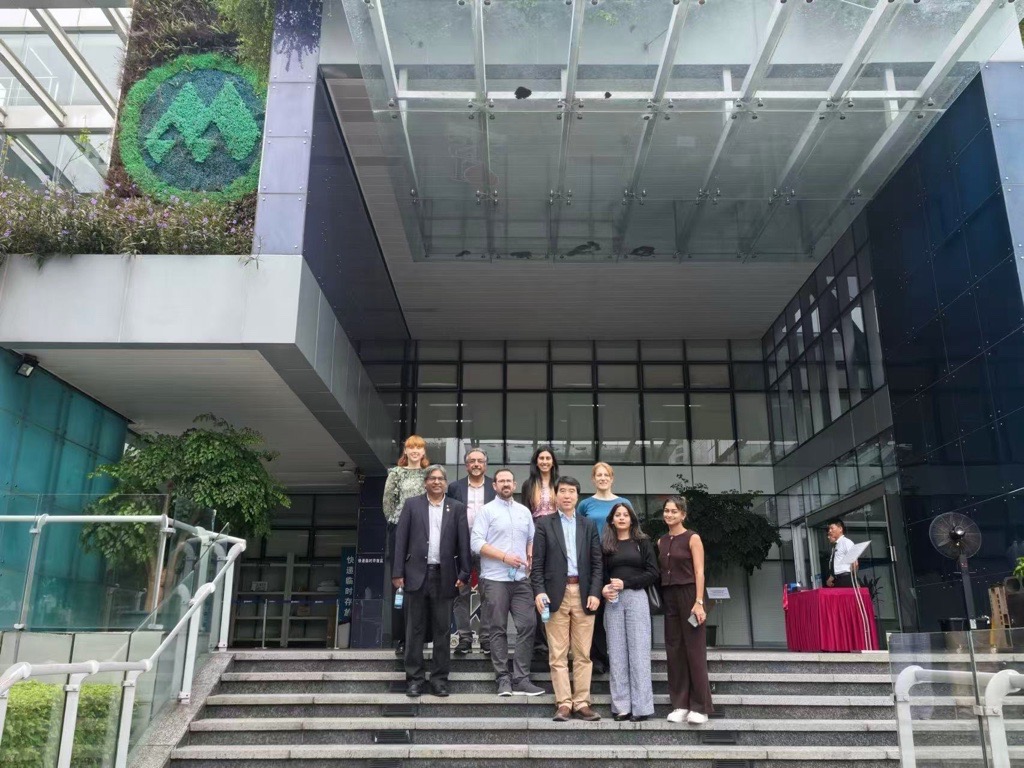Nine people pose on the steps outside a modern office building with a glass entrance and green wall feature.