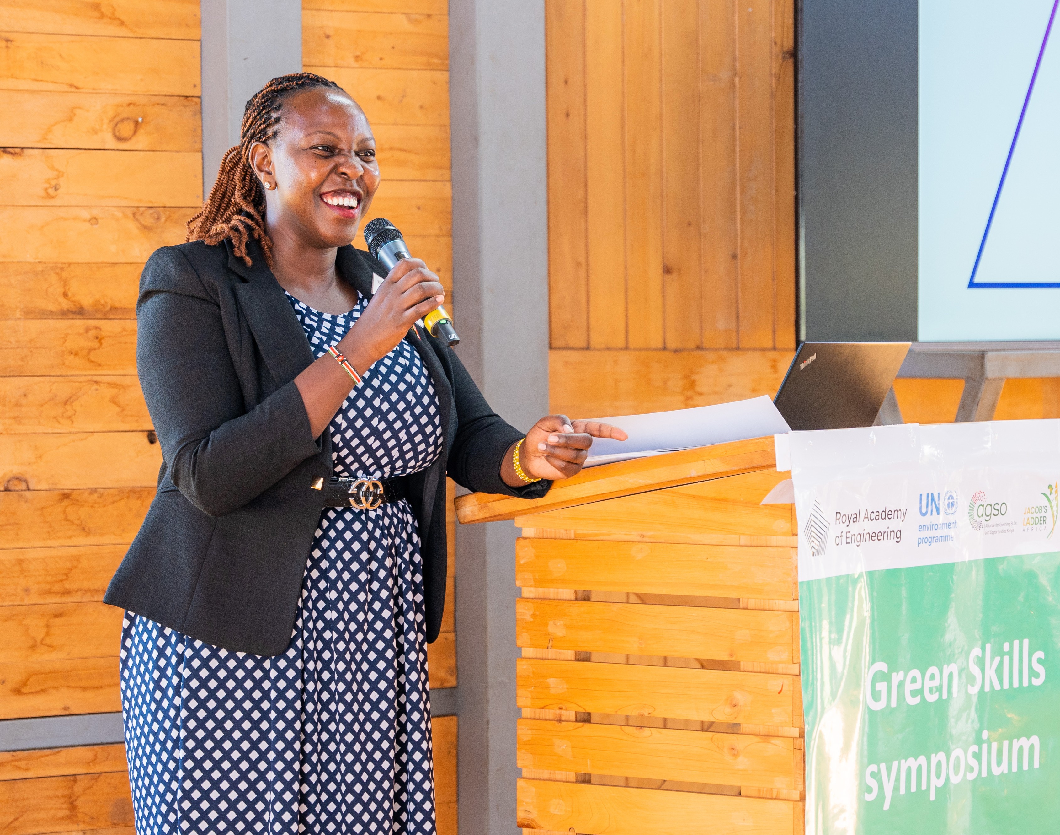 A black woman in a polkadot dress and black blazer stands next to a podium holding a microphone.