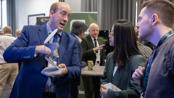 Attendees talking at a conference exhibition. One person holds an item and is showing the other people.