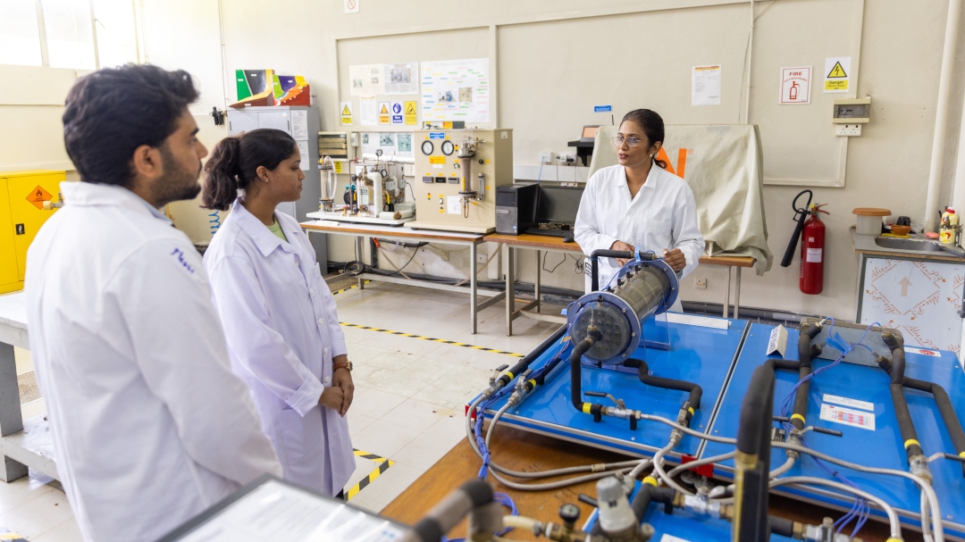 An Indian woman lecturer, dressed in a white lab coat, explains how to use some of the equipment in a university classroom. She is speaking to a young Indian male and a young Indian female student, both also wearing white lab coats. The classroom has tables with equipment, safety tape on the floors, and posters and safety notices on the walls.