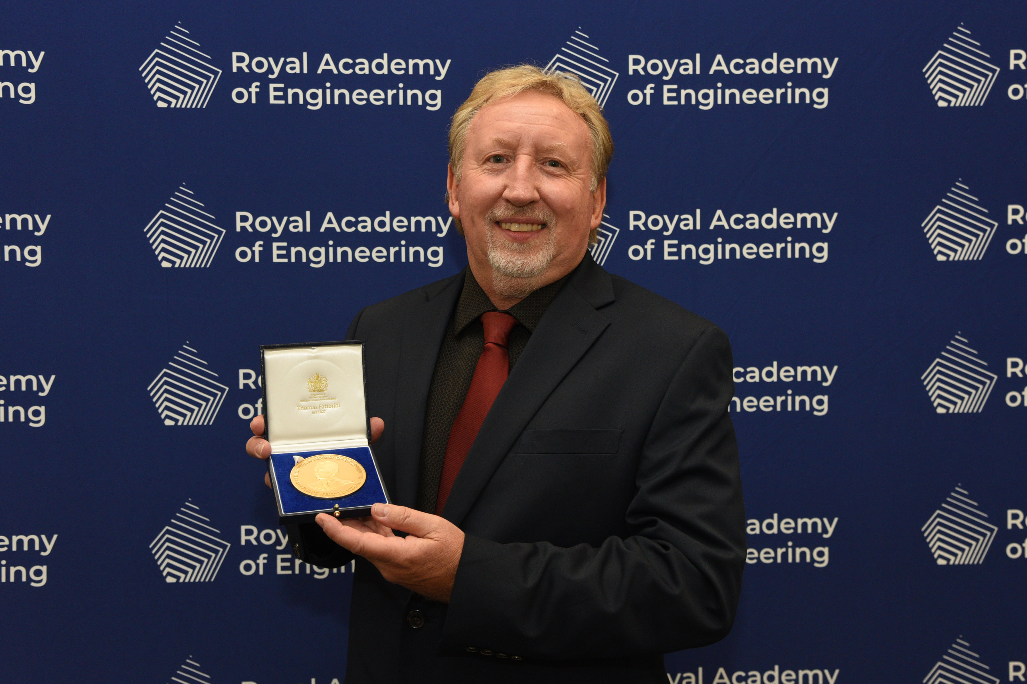Professor Graham Reed FREng holding his Sir Frank Whittle Medal in front of a blue screen with the Royal Academy of Engineering logo
