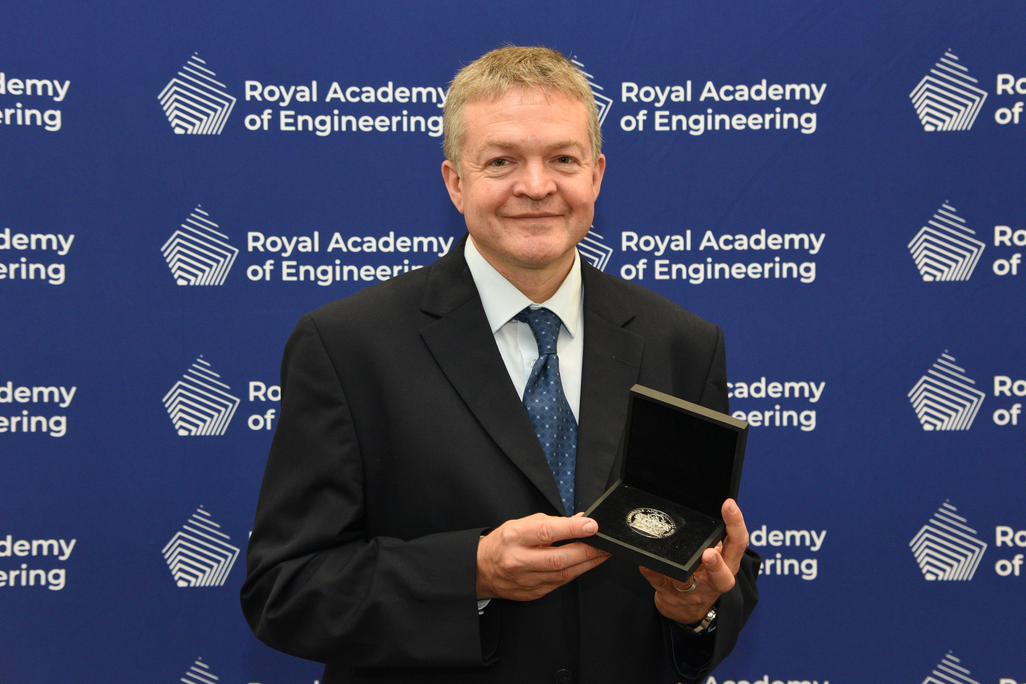 Dr Robert Leah holding his RAEng Armourers and Brasiers Prize in front of a blue screen with the Royal Academy of Engineering logo