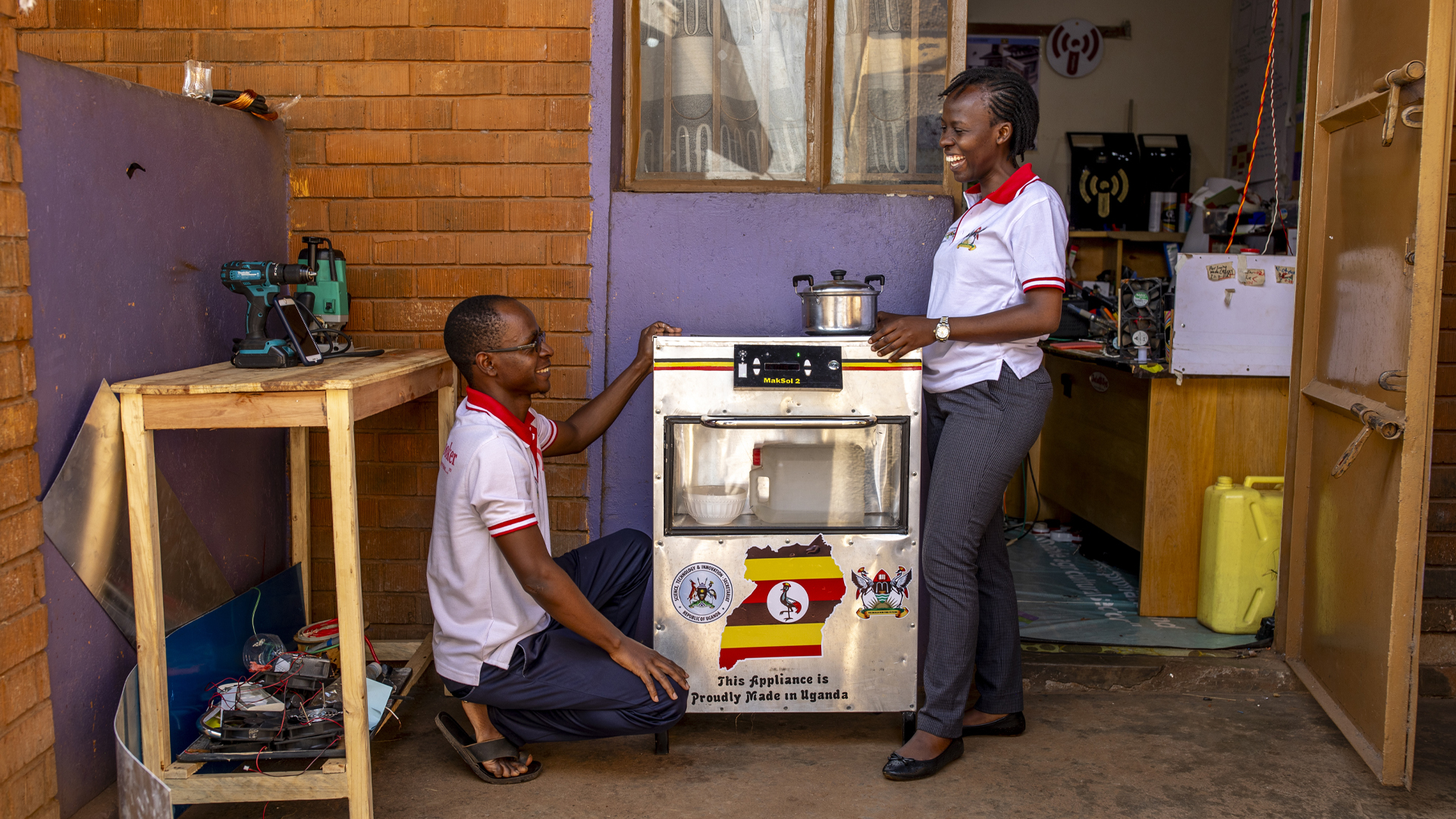 A smiling, Black man and woman wearing matching shirts look at one another over a metal stove, which is branded with the Ugandan flag. The man is kneeling on the ground while the woman is standing. The photo is taken outside, in front of a building, with tools visible on the inside.