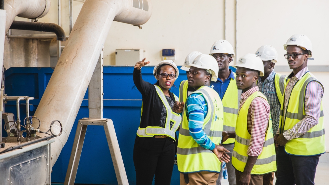 A black woman wearing glasses, a white hard hat and a reflective vest stands in an industrial setting, gesturing towards something in the foreground. Six young black men wearing hard hats and reflective vests over collared shirts look attentively towards where she is pointing. In the background are the white and blue walls of a factory, and a large industrial pipe runs alongside the group