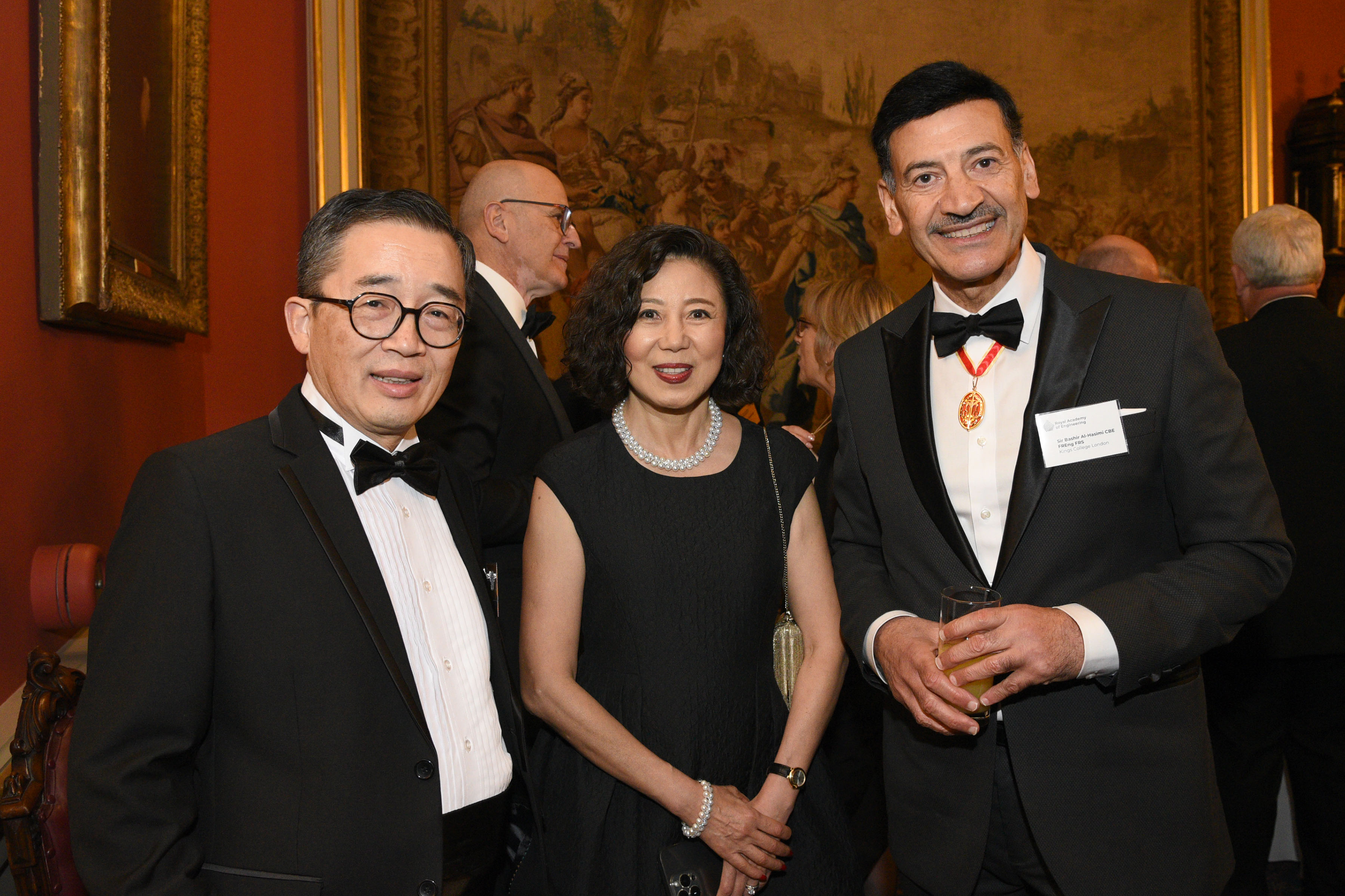 Three engineers wearing black tie, standing in front of a large oil painting in a grand room.