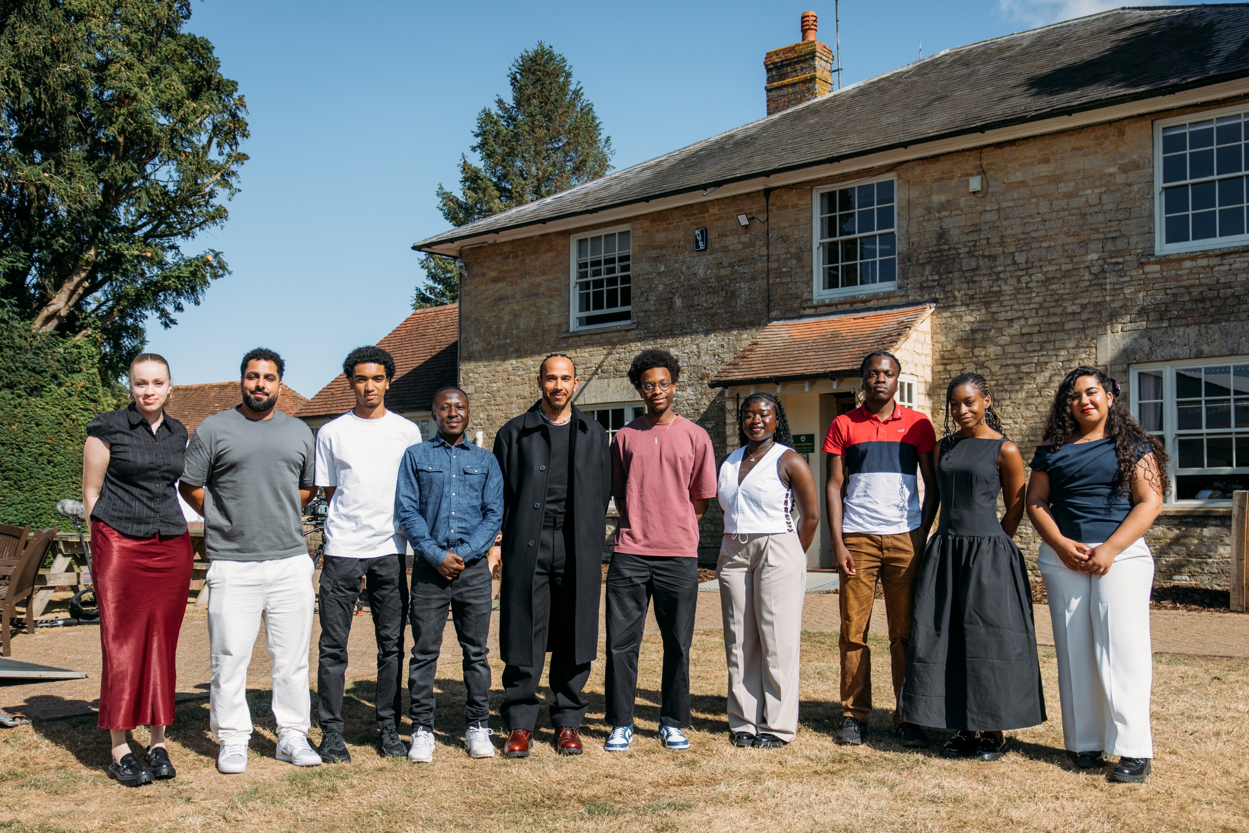 A group of young people posing for a group photo with Sir Lewis Hamilton outside on a sunny day.