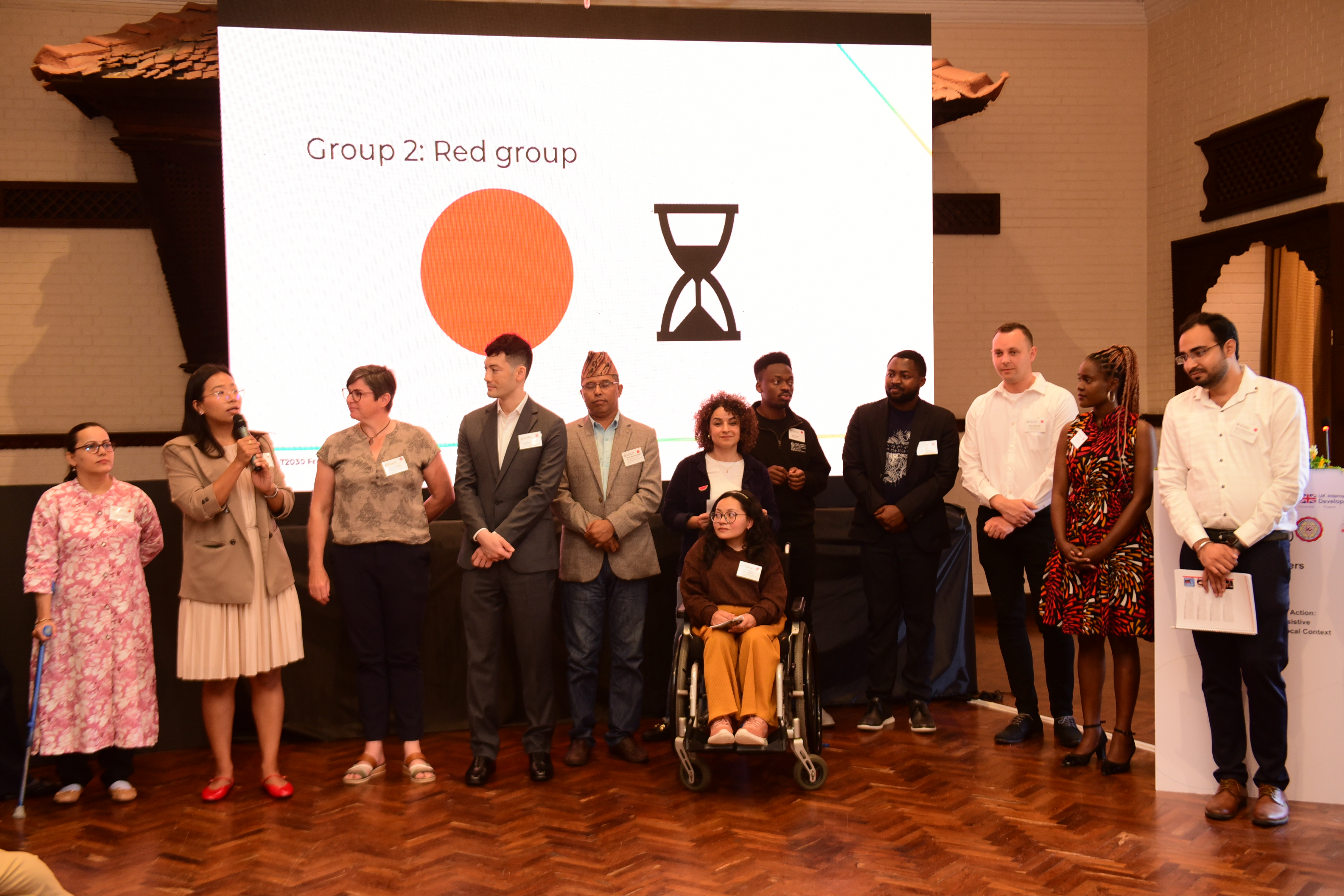 A line of event participants is at the front of a room, including one person using a wheelchair. The scene shows a group activity that may require on-the-go adjustments to spacing, audiovisual factors, visibility, and facilitation, to make sure all participants can take part comfortably.