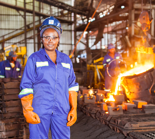 A Black woman in blue overalls, orange safety gloves, a blue hardhat and safety goggles, faces the camera and smiles. Behind her in the blurred background is a foundry where another person in blue overalls and a safety hat is turning a wheel that tips a large drum of glowing, molten metal into moulds.